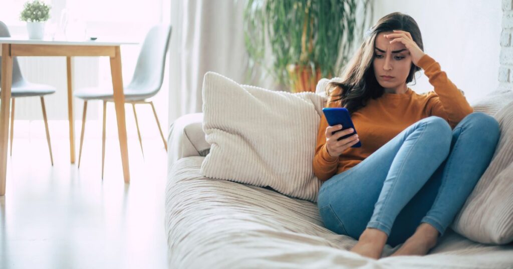 ソファに寄りかかり不安そうな顔でスマホを見つめる女性