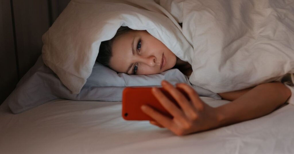 消灯した部屋のベッドに寝そべりスマホをチェックする女性